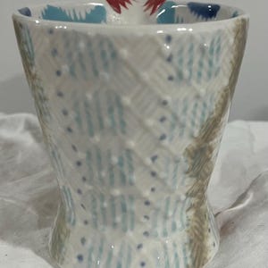 May include: A handmade ceramic cup with a unique, textured design. The cup features a white base with blue and brown patterns, and small blue dots. The rim has a blue and red design.