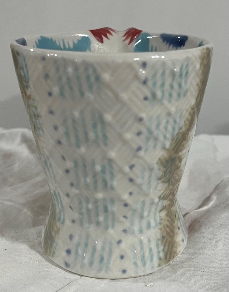 May include: A handmade ceramic cup with a unique, textured design. The cup features a white base with blue and brown patterns, and small blue dots. The rim has a blue and red design.