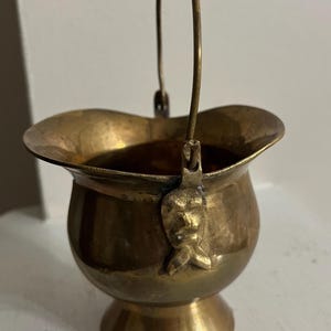 May include: A small, brass bucket with a handle. The bucket has a decorative design on the side and a flared lip.