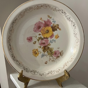 May include: A white ceramic serving dish with a gold rim and floral design. The dish features a bouquet of pink, yellow, and blue flowers in the center. A gold decorative pattern encircles the rim. The dish is supported by a gold-colored metal stand.