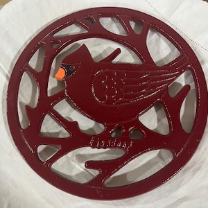 May include: A circular, deep red cast iron trivet featuring an openwork design of a cardinal bird perched on a branch. The bird has a bright orange beak and a black face mask, with the entire piece showcasing a detailed, textured metal finish.