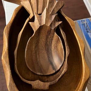 May include: A set of wooden bowls in the shape of a pineapple. The bowls are nested inside each other, with the largest bowl forming the outer shape and the smallest bowl at the center. The wood has a warm, natural tone.