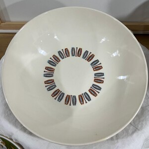 May include: A large, round, off-white ceramic bowl featuring a central circular pattern composed of repeating, stylized geometric shapes in alternating shades of burnt orange and muted blue, arranged in a ring around the interior base of the bowl.