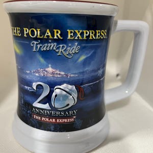 May include: A white ceramic mug featuring a winter scene with the text "THE POLAR EXPRESS Train Ride" and "20th ANNIVERSARY THE POLAR EXPRESS". The mug has a white handle and a white base with a red rim.