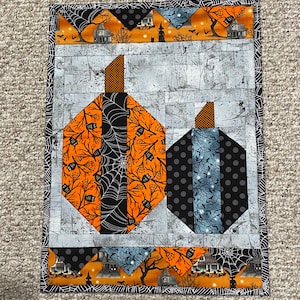 May include: A Halloween-themed quilted wall hanging with two pumpkins. The larger pumpkin is orange and black with a spiderweb pattern. The smaller pumpkin is blue and black with white polka dots. Both pumpkins are surrounded by a black and white spiderweb border.