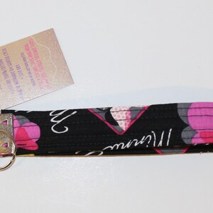 Puede incluir: Un llavero de mu&ntilde;eca negro con un estampado floral en rosa, gris y amarillo. El llavero tiene un cierre de metal plateado y una etiqueta adjunta. La etiqueta tiene texto impreso.
