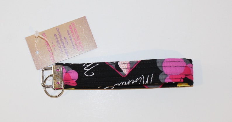 Puede incluir: Un llavero de mu&ntilde;eca negro con un estampado floral en rosa, gris y amarillo. El llavero tiene un cierre de metal plateado y una etiqueta adjunta. La etiqueta tiene texto impreso.