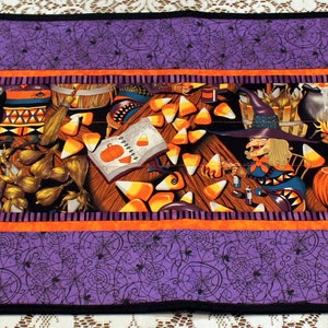 May include: A purple and orange Halloween table runner with a witch, cauldron, and candy corn design. The runner has a black border and a purple background with a spiderweb pattern.