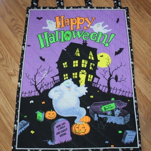May include: Purple quilted wall hanging with a black house, a ghost, and a graveyard. The text "Happy Halloween!" is in green and orange letters.