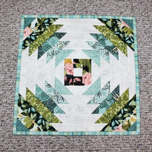 May include: A square quilt with a pineapple design. The quilt features a white background with a central square of floral fabric. Green, teal, and floral patterned triangles radiate outwards, framed by a teal and white border.