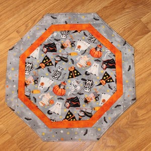 May include: Octagonal Halloween-themed table runner. The center features a gray fabric with Halloween characters like ghosts, cats, and pumpkins. An orange border frames the center, and the outer edge is gray with stars and bats.
