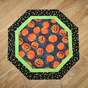 May include: Octagonal table runner with a black background and a green border. The center features a repeating pattern of orange jack-o'-lanterns with black eyes and mouths, and spiderwebs.
