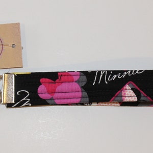 Puede incluir: Llavero de pulsera de tela negra con un dise&ntilde;o de Minnie Mouse en rosa y blanco. El llavero tiene un cierre de metal plateado y un llavero. Una peque&ntilde;a etiqueta est&aacute; adjunta con el texto "Totally Toile" y "Handmade Designs by Lyn Lindsey Sky".