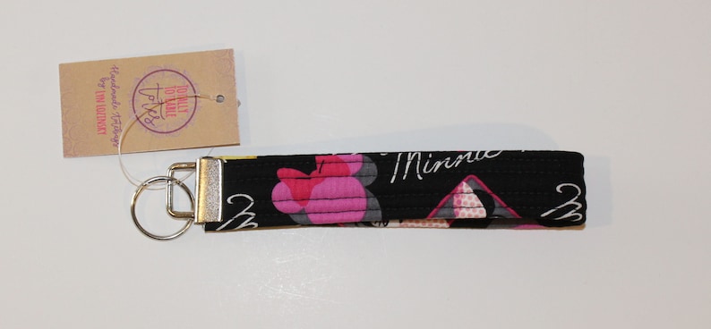 Puede incluir: Llavero de pulsera de tela negra con un dise&ntilde;o de Minnie Mouse en rosa y blanco. El llavero tiene un cierre de metal plateado y un llavero. Una peque&ntilde;a etiqueta est&aacute; adjunta con el texto "Totally Toile" y "Handmade Designs by Lyn Lindsey Sky".