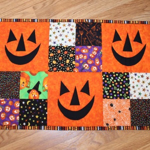 May include: A Halloween-themed table runner with a patchwork design featuring orange, black, green, and purple fabric squares. The squares have various patterns, including pumpkins, stars, and spiderwebs. Three black jack-o'-lantern faces are appliqued onto the orange squares.