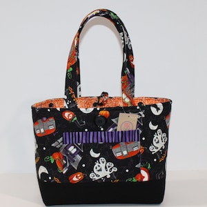 May include: Black tote bag with a Halloween theme. The bag features a variety of spooky designs, including ghosts, pumpkins, and haunted houses. The bag has a purple and white striped pocket and a black button.