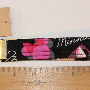 Puede incluir: Llavero de tela negra con un cierre y un llavero de metal dorado. La tela presenta un estampado floral rosa y la palabra "Minnie" en escritura blanca. Se adjunta una etiqueta con texto. El llavero mide unos 15 cm de largo.