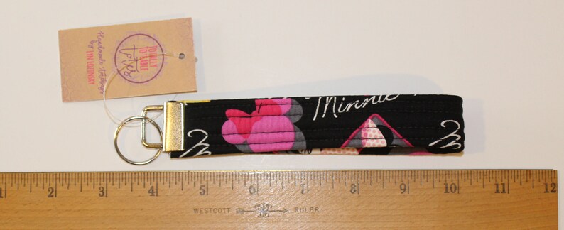 Puede incluir: Llavero de tela negra con un cierre y un llavero de metal dorado. La tela presenta un estampado floral rosa y la palabra "Minnie" en escritura blanca. Se adjunta una etiqueta con texto. El llavero mide unos 15 cm de largo.