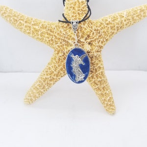 Puede incluir: Collar con colgante plateado con un diseño ovalado en tonos azules y una figura de ángel plateada. El collar, expuesto sobre una estrella de mar, tiene un cordón negro. La dirección del sitio web es visible.