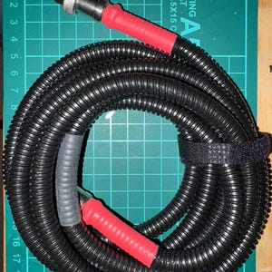 May include: A black coiled cable with red insulated ends and a silver connector. The cable is wrapped with black tape and secured with a black velcro strap. The cable is on a green cutting mat with a grid pattern and the text "CUTTING MAT 22.5X15 C".