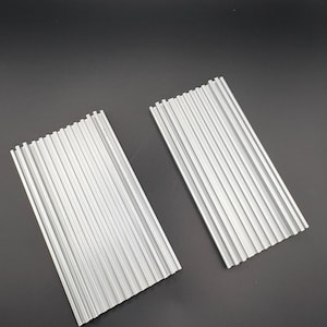 Puede incluir: Dos piezas metálicas plateadas y estriadas se muestran sobre un fondo oscuro. Las piezas rectangulares tienen estrías paralelas a lo largo de su longitud. El metal tiene un acabado mate.