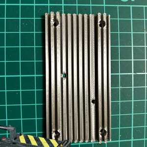 Puede incluir: Un disipador de calor de metal plateado con múltiples aletas y varios agujeros. El disipador de calor está sobre una superficie de rejilla verde.