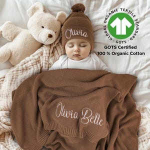 Peut inclure: Une couverture en tricot marron pour bébé avec le nom "Olivia Belle" brodé dessus. Un bébé portant un bonnet en tricot marron avec le nom "Olivia" brodé dessus dort sur la couverture. La couverture est certifiée GOTS et fabriquée en coton biologique 100%.