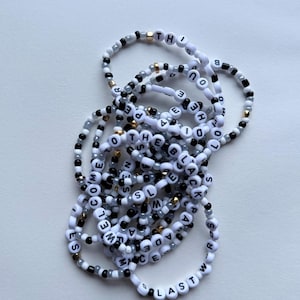 May include: A collection of beaded bracelets with white letter beads spelling out phrases like "THE BLACK" and "WELCOME". The bracelets feature black, gray, and gold accent beads. The bracelets are on a white surface.