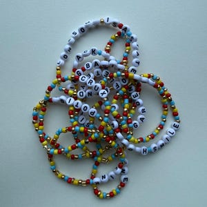 May include: Several beaded bracelets with white letter beads spelling out words. The bracelets feature red, yellow, blue, and gold beads. The bracelets are arranged in a tangled pile. The bracelets are made of small beads.