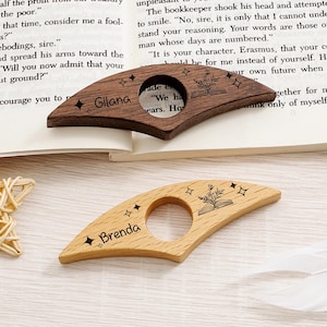 May include: Two wooden page holders, one dark brown and one light brown, rest on an open book. The dark brown holder is engraved with the name "Gilana" and a floral book design. The light brown holder is engraved with the name "Brenda".