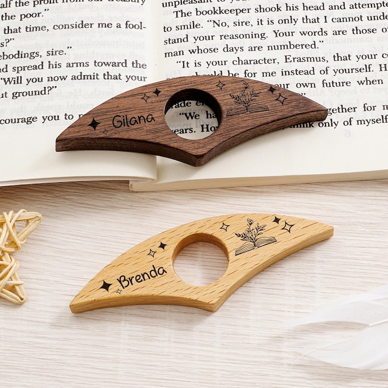 May include: Two wooden page holders, one dark brown and one light brown, rest on an open book. The dark brown holder is engraved with the name "Gilana" and a floral book design. The light brown holder is engraved with the name "Brenda".