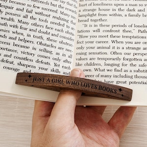 May include: A wooden bookmark with the text "JUST A GIRL WHO LOVES BOOKS" engraved on it. The bookmark is dark brown and is placed on the pages of a book. The text is in a simple, readable font.