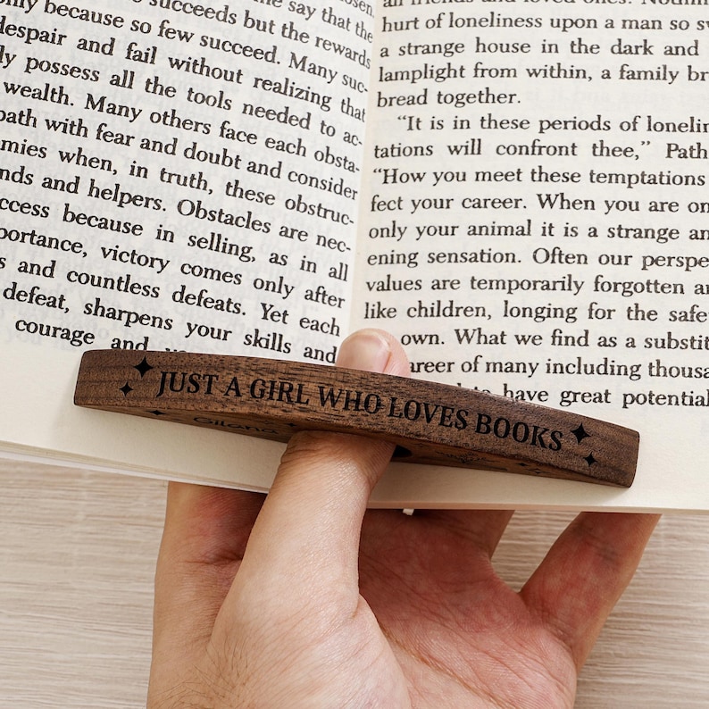 May include: A wooden bookmark with the text "JUST A GIRL WHO LOVES BOOKS" engraved on it. The bookmark is dark brown and is placed on the pages of a book. The text is in a simple, readable font.