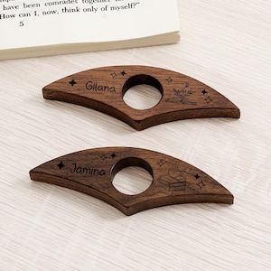 May include: Two wooden book page holders with a dark brown finish. Each has a circular opening and is engraved with a name, stars, and a book-themed design. One says "Gilana" and the other says "Jamina".
