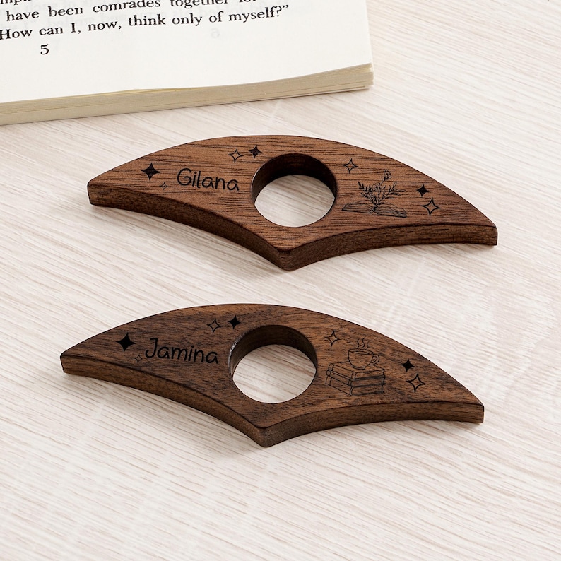 May include: Two wooden book page holders with a dark brown finish. Each has a circular opening and is engraved with a name, stars, and a book-themed design. One says "Gilana" and the other says "Jamina".