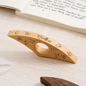 Puede incluir: Un sujetapáginas de madera con un diseño curvo y un agujero para el dedo. El soporte está grabado con el nombre "Heather" y diseños florales y de mariposas. El soporte está hecho de madera clara.