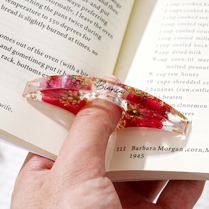 May include: A clear resin book page holder with red floral inclusions and gold flakes. The holder has the name "Bianca" in script and butterfly designs. It is designed to slip over a finger to hold a book open.
