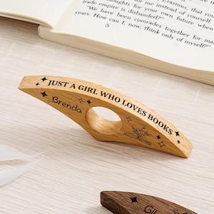 May include: A wooden book page holder with the text "JUST A GIRL WHO LOVES BOOKS" and the name "Brenda". The holder is light brown with a circular opening and star and book illustrations.
