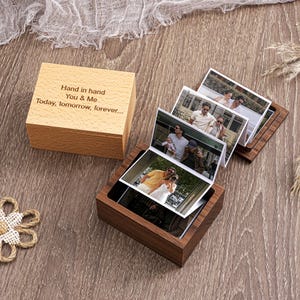 May include: A wooden photo box with a hinged lid and a dark wood base. The lid is engraved with the text "Hand in hand, You & Me, Today, tomorrow, forever...". The box contains several small photos. The box is on a wooden surface.