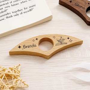 May include: Wooden book holder with a circular opening and engraved text. The light-colored wood features the name "Brenda" and star designs. A second holder is visible with the name "Gilana". The design includes an open book and floral elements.