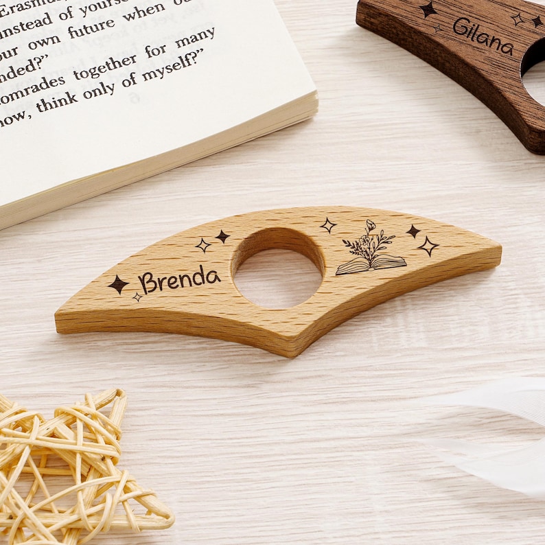 May include: Wooden book holder with a circular opening and engraved text. The light-colored wood features the name "Brenda" and star designs. A second holder is visible with the name "Gilana". The design includes an open book and floral elements.