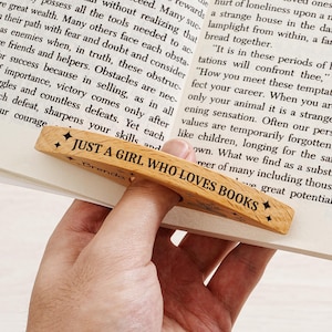 May include: Wooden bookmark with the text "JUST A GIRL WHO LOVES BOOKS" and star accents. The bookmark is made of light-colored wood and is designed to slip over a page in a book.