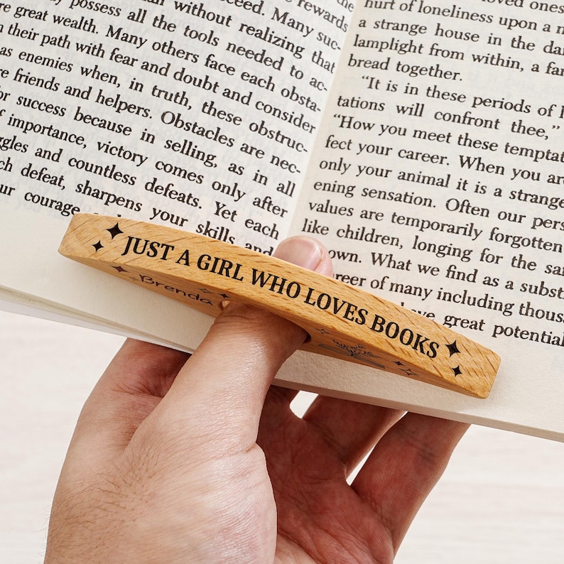 May include: Wooden bookmark with the text "JUST A GIRL WHO LOVES BOOKS" and star accents. The bookmark is made of light-colored wood and is designed to slip over a page in a book.