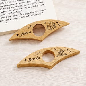 May include: Two wooden book page holders with a cutout circle for the thumb. One is engraved with "Halona" and the other with "Brenda". Both have star and book illustrations. The book page holders are on a light wood surface.