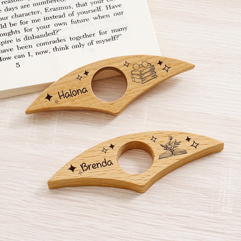 May include: Two wooden book page holders with a cutout circle for the thumb. One is engraved with "Halona" and the other with "Brenda". Both have star and book illustrations. The book page holders are light brown.