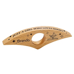 May include: A wooden book holder with the text "JUST A GIRL WHO LOVES BOOKS" and the name "Brenda" engraved on it. The holder features a circular opening for the finger and a design of an open book with a plant. Decorative star shapes are also engraved.