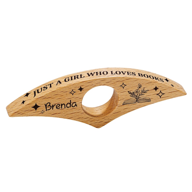 May include: A wooden book holder with the text "JUST A GIRL WHO LOVES BOOKS" and the name "Brenda" engraved on it. The holder features a circular opening for the finger and a design of an open book with a plant. Decorative star shapes are also engraved.