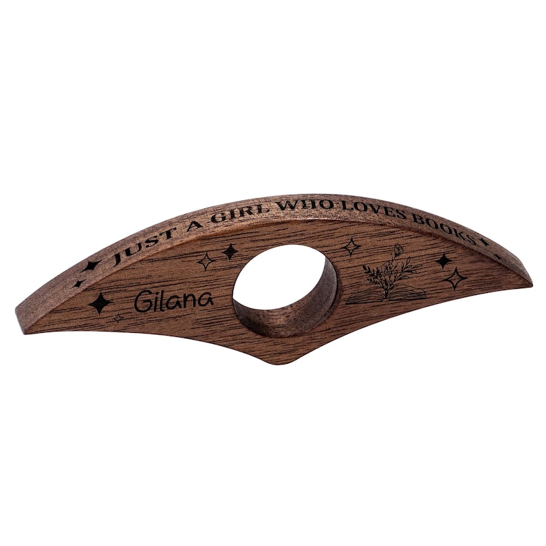 May include: A wooden book page holder with a circular opening and a curved design. The text "JUST A GIRL WHO LOVES BOOKS" is engraved on the top, along with the name "Gilana" and star and plant designs. The holder is made of dark brown wood.