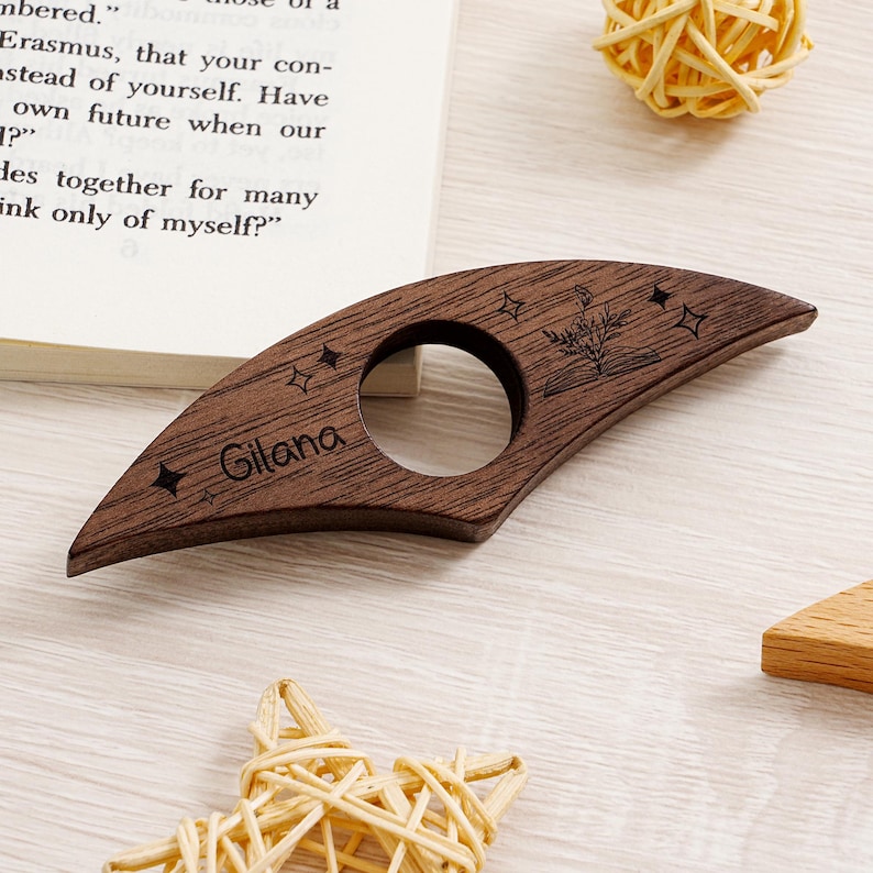 May include: A dark brown wooden thumb page holder with a circular opening and engraved stars and floral design. The name "Gilana" is engraved on the holder. The thumb page holder is designed to hold a book open.