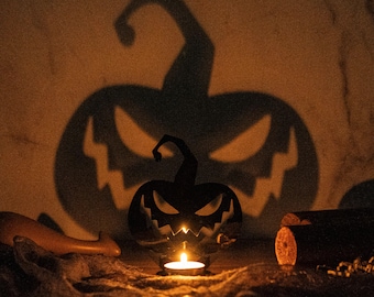May include: A black metal pumpkin candle holder with a carved jack-o'-lantern face. The candle holder is lit with a small candle and casts a shadow of a larger pumpkin on the wall behind it.